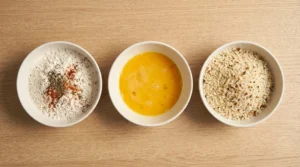Three bowls arranged in a row containing flour with spices, beaten eggs, and panko breadcrumbs, demonstrating the setup for crispy chicken tenders without deep frying.