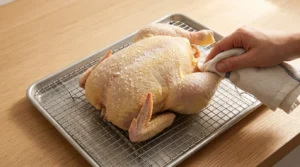 A hand patting dry a whole chicken on a wire rack set over a sheet pan, prepared for Garlic Herb Roast Chicken.