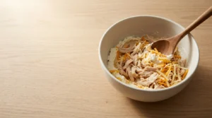 Softened cream cheese, shredded rotisserie chicken, and shredded cheese being mixed in a bowl with a wooden spoon, prepared for Halloween Chicken Mummy Wraps.