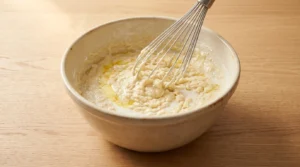A ceramic bowl containing dumpling batter being mixed with a wire whisk, prepared for slow cooker chicken and dumplings