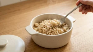 A hand using a fork to fluff freshly cooked brown rice or quinoa in a ceramic bowl on a light wood countertop, prepared for high protein chicken bowl meal prep.